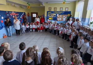 w szatni przedszkolnej zgromadzone dzieci i nauczycielki, wszyscy ubrani na galowo, dzieci mają biało-czerwone wianki , przygotowują siędo wspólnego śpiewania hymnu