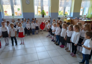 w szatni przedszkolnej zgromadzone dzieci i nauczycielki, wszyscy ubrani na galowo, dzieci mają biało-czerwone wianki , przygotowują siędo wspólnego śpiewania hymnu