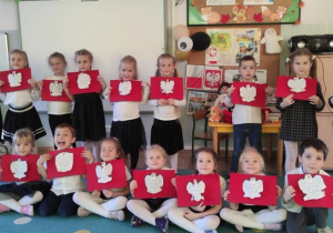 grupa dzieci ubranych odświętnie pozuje do zdjęcia z pracami wykonanymi na zajęciu plastycznym, Jest to biały orzeł na czerwonym tle