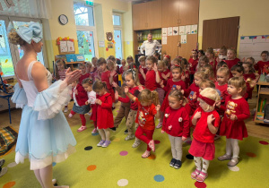 przedszkolaki zgromadzone w klasie, stoją a naprzeciwko nich jest artystka, która pokazuje ruchy taneczne, a dzieci powtarzają