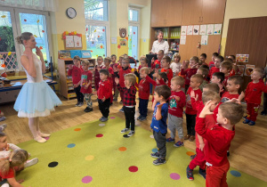 przedszkolaki zgromadzone w klasie, stoją a naprzeciwko nich jest artystka, która pokazuje ruchy taneczne, a dzieci powtarzają