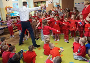 przedszkolaki zgromadzone w klasie, stoją a naprzeciwko nich jest organizator koncertu, który pokazuje ruchy taneczne, a dzieci powtarzają