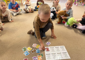grupa dzieci siedzi na dywanie w klasie, jeden z chłopców jest na środku i wykonuje zadanie z misiami