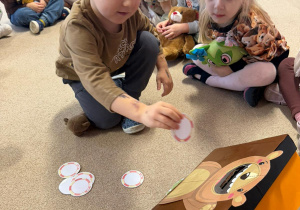 grupa dzieci siedzi na dywanie w klasie, jeden z chłopców jest na środku i wykonuje zadanie z misiami