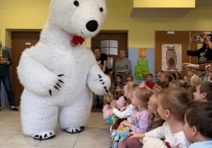 dzieci siedząna ławkach w satni, naprzeciwko nich biały wielki miś, który podchodzi do nich i wita się