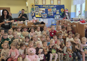 duża grupa dzieci z misiami w dłoniach siedzi na ławkach w szatni, uczestniczą w spotkaniu z okazji Dnia Misia, obok stoją panie