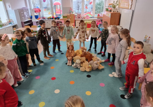 grupa dzieci stoi w kole trzymają sięza ręce, w środku kołą siedzi dziewczynka, obok niej leżą pluszaki