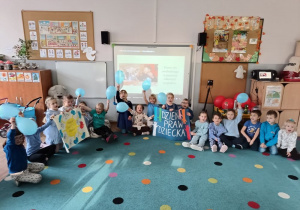 grupa dzieci siedzi na wywanie, ubrane na niebiesko, niektóre mają w rękach niebieskie balony, inne trzymają plakaty zrobione przez siebie nawiązujące do tematu dnia