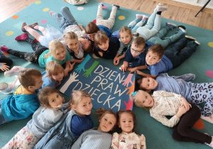 dzieci ubrane na niebiesko leżą na dywanie , pośrodku leży plakat z napisem "dzień praw dziecka"