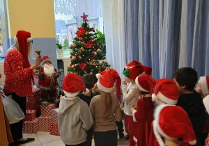 grupa dzieci w przedszkolnej szatni, ubrane na czerwono, niektóre mają mikołajkowe i świąteczne elementy ubioru, gromadząsięobok choinki przy której jest pani dyrektor dzwoniąca dzwonkiem