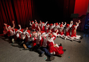 dzieci w mikołajowych strojach na scenie w MOK, siedzą w dwóch rzędach, mają uniesione ręce