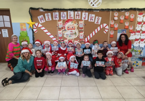 grupa dziec i nauczycielki pozują do zdjęcia na tle mikołajkowej dekoracji w szatni, wszystcy ubrani na czerwono, niektórzy w mikołajowych czapkach