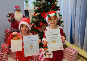 dwie dziewczynki przy choince, w rękach mają dyplony za udział w konkursie światecznym