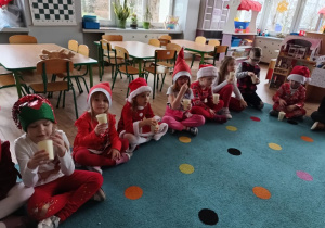 grupa dzieci siedzui na dywanie, ubrane na czerwono, w rekach trzymają kubeczki z popcornem