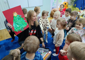 grupa dzieci w bibliotece, zgromadzone wkół pani bibliotekarki, która pokazuje im pracę plastyczną - choinkę