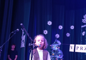 dziewczynka stoi na scenie, za nią zimowa dekoracja, dziecko śpiewa