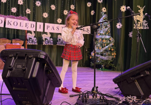 dziewczynka stoi na scenie, za nią zimowa dekoracja, dziecko śpiewa