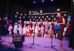 grupa dzieci i nauczycielki stoją na scenie, jedna z pań stoi przy mikrofonie i mówi