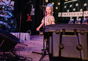 dziewczynka stoi na scenie, za nią zimowa dekoracja, dziecko śpiewa