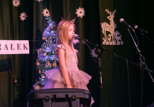 dziewczynka stoi na scenie, za nią zimowa dekoracja, dziecko śpiewa