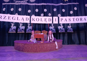 dwie dziewczynki siedzą na scenie, za nimi zimowa dekoracja