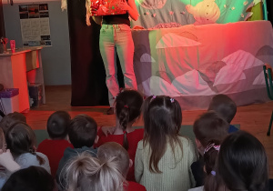 dzieci siedzą na dywanie oglądają przedstawienie, przed mini scenografia i aktorka