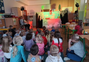 dzieci siedzą na dywanie oglądają przedstawienie, przed mini scenografia, z lewej strony siedzi nauczycielka
