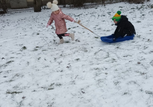 jedno dziecko ciągnie po śniegu kolegę , który siedzi na sankach