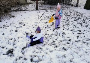 dwie dziewczynki podczas zabaw na śniegu , jedna siedzi na ziemi, druga stoi z "jabłuszkiem" do zjazdów w ręce
