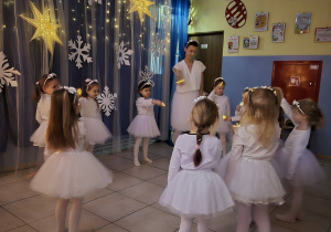 dziewczynki w strojach aniołków wykonują taniec na występie, obok nauczycielka ubrana na biało