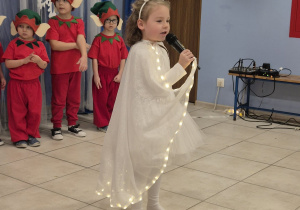dziewczynka ubrana na biało śpiewa, w tle chłopcy przebrani za elfy