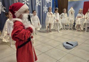 grupa dzieci prezentuje występ świateczny rodzicom,na pierwszym planie chłopiec przebrany za św. Mikołaja, w tle dziewczynki ubrane na biało