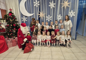 grupa dzieci i nauczycielka pozujądo zdjęcia z Mikołajem, w tle świateczna dekoracja