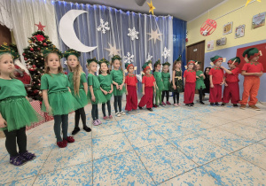 grupa dzieci przebranych za choineczki i elfy podczas występu światecznego, w tle zimowa dekoracja