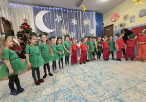 grupa dzieci przebranych za choineczki i elfy podczas występu światecznego, w tle zimowa dekoracja