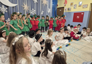 grupa dzieci prezentuje się przed rodzicami na światecznym występie, niektóre stoją, niektóre siedzą na podłodze i będą grać na dzwonkach