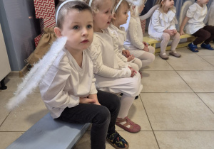 grupa dzieci przebranych za aniołki siedzi na ławeczce podczas występu