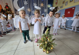 grupa dzieci , niektóre przebrane za aniołki, pozostałe elegancko ubrane prezentuje rodzicom świąteczne przedstawienie, na pierwszym planie czworo dzieci, przed nimi stoi mała choineczka