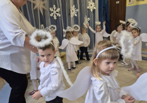 grupa dzieci , niektóre przebrane za aniołki, pozostałe elegancko ubrane prezentuje rodzicom świąteczny taniec , obok jest nauczycielka