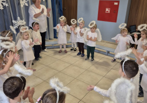 grupa dzieci , niektóre przebrane za aniołki, pozostałe elegancko ubrane prezentuje rodzicom świąteczny taniec , z nimi stoją ich panie