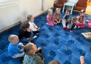 dzieci siedzą na dywanie słuchają pani bibliotekarki, obok leżą książki