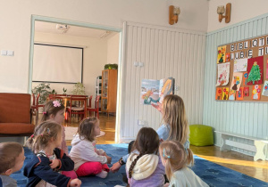 dzieci siedzą na dywanie słuchają opowiadań czytanych przez bibliotekarkę