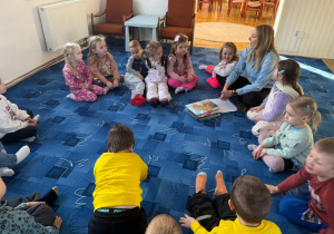 dzieci siedzą na dywanie słuchają pani bibliotekarki, obok niej leżą książki