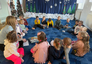 dzieci siedzą na dywanie słuchają pani bibliotekarki, obok niej leżą książki