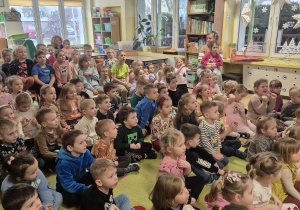 przedszkolaki zgromadzone w sali, siedzą na podłodze oglądają występ muzyczny