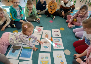 grupa dzieci siedzi na dywanie przed nimi plansze edukacyjne i wykonane przez nie zadania projektowe