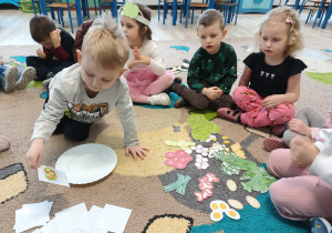 grupa dzieci siedzi na dywanie, jedno z nich wykonuje zadanie z obrazkami warzyw