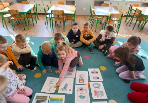 grupa dzieci siedzi na dywanie przed nimi plansze edukacyjne i wykonane przez nie zadania projektowe