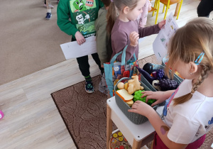 dzieci bawią się w zabawę fabularyzowaną - stojąw kolejce do sklepu, niektóre mają w rękach koszyki na zakupy