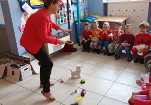 Nauczycielka prowadzi tematyczne zajęcia. W tle na ławeczkach siedzą dzieci.